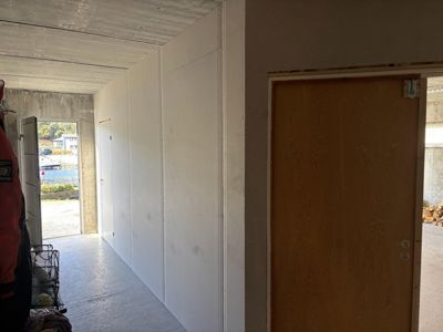 Fotografi av en gang eller korridor i en bygning under oppføring eller renovering. Bildet viser en betonggulv, hvite panelvegger, en åpen dør og utsikt ut til vann og nabolag.