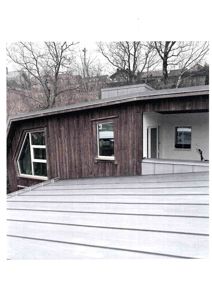 Fotografi av en bygning sett fra siden, som viser fasaden med mørk trepaneling, vinduer og en del av taket i forgrunnen.