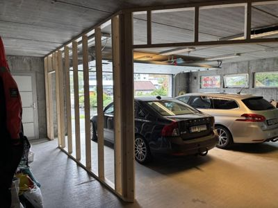 Fotografi av en garasje eller underetasje under ombygging, med en ny oppstilt tømmerkonstruksjon (rammeverk) for en innvendig vegggang eller romdeling. Det er parkert to biler i bakgrunnen.