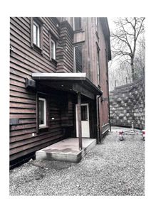 Fotografi av en bygning sett fra siden, som viser fasade, vinduer, en utvendig dør med tak, og omgivelser med grus og en steinmur.