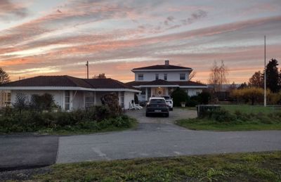 Fotografi av en bolig med tilhørende bygninger, sett fra veien mot solnedgang.