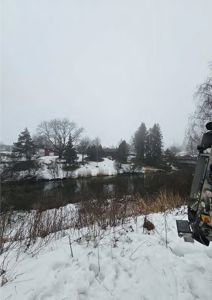Et fotografi av en snødekt landskap med en elv, trær og en bygning i bakgrunnen.