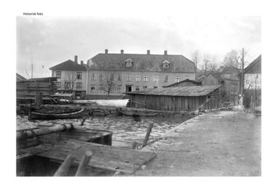 Et historisk svart-hvitt fotografi som viser en bygning ved vannet med tilhørende mindre bygninger og båter.