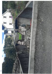 Fotografi av en eksisterende bygning (hvit villa) sett fra siden, med en betongmur i forgrunnen og en grusvei.