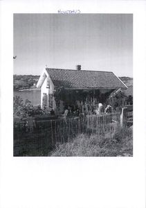 Sort-hvitt fotografi av en liten hytte eller hovedhus ved vannet, med hage og omgivelser.