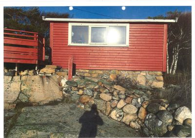 Fotografi av en rød hytte sett fra siden, med en steinmur i forgrunnen og et rødt veranda- eller terrasseelement til venstre.