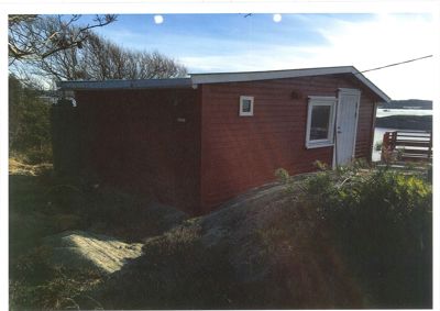 Fotografi av en rød hytte eller liten bygning sett fra siden, med omgivende natur og vann i bakgrunnen.