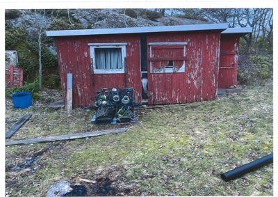 Fotografi av en rød, utvasket uthusbygning (kanskje fiskehytte eller båthavn) med fiskekroker foran, sett fra en vinkel som viser fasade og deler av omgivelsene.