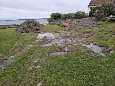 Fotografi av et utendørs område med grøfting, to store jordhauger, en steinmur og en del av en bygning i bakgrunnen.