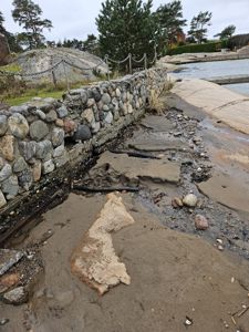 Fotografi av en steinmur ved en vei eller gangvei, med en svart slange eller kabel som ligger i grøfta ved siden av muren.