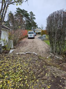 Fotografi av en tomt med en bil parkert på en grusvei, en bygning i bakgrunnen og trær.