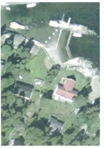 Satellittbilde eller flyfoto som viser en tomt med flere bygninger, en kai og vann.