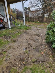 Fotografi av en bakgård med en gul hytte i venstre side, en grusete bakgård, og en hekk i bakgrunnen.