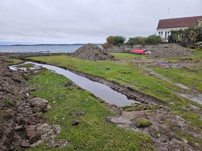 Fotografi av et bygge- eller anleggsområde ved vannet med grus, en grøft og en bygning i bakgrunnen.