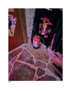 Fotografi av en innendørs hjørne med en brannslukker, en steinlagt gulvflate og en t-skjorte hengende på veggen.