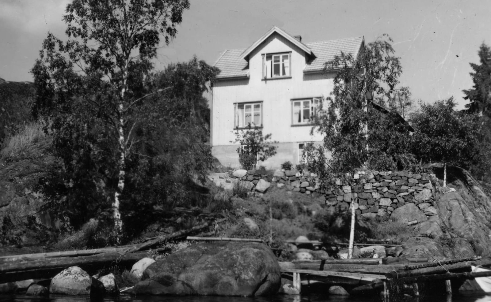 Sort-hvitt fotografi av en hvit villa med etasjehus, plassert på en steinete bakke ved vannet.