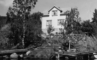 Sort-hvitt fotografi av en hvit villa med etasjehus, plassert på en steinete bakke ved vannet.