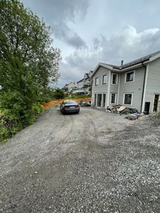 Fotografi av en grå bebygd tomt med en nybygd eller renovert villa i høyre side, en bil på en grusvei og naboer i bakgrunnen.