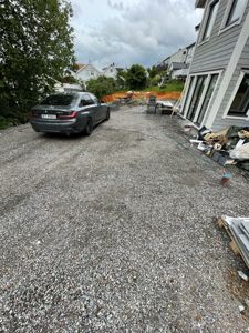 Fotografi av en grusplass foran en grå villa under ombygging eller renovering, med en bil parkert og byggematerialer i bakgrunnen.