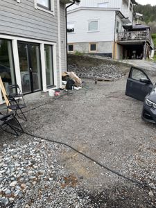 Fotografi av en byggeplass eller tomt med en grå fasadetett bygning i forgrunnen, en annen hvit bygning i bakgrunnen, og en bil med åpen dør på høyre side.
