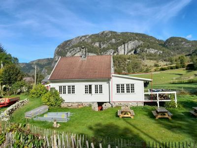 Fotografi av en hvit, trepanelt hytte med rødt tak, sett fra siden. Bildet viser også omgivelser som en grønn hage, en steinmur, et terrasseområde, og en stor fjellvegg i bakgrunnen.