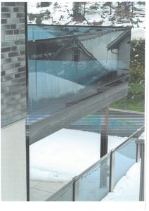 Fotografi av en moderne balkong eller terrasse med glassrailing, snø på bakken og en steinmur til venstre.