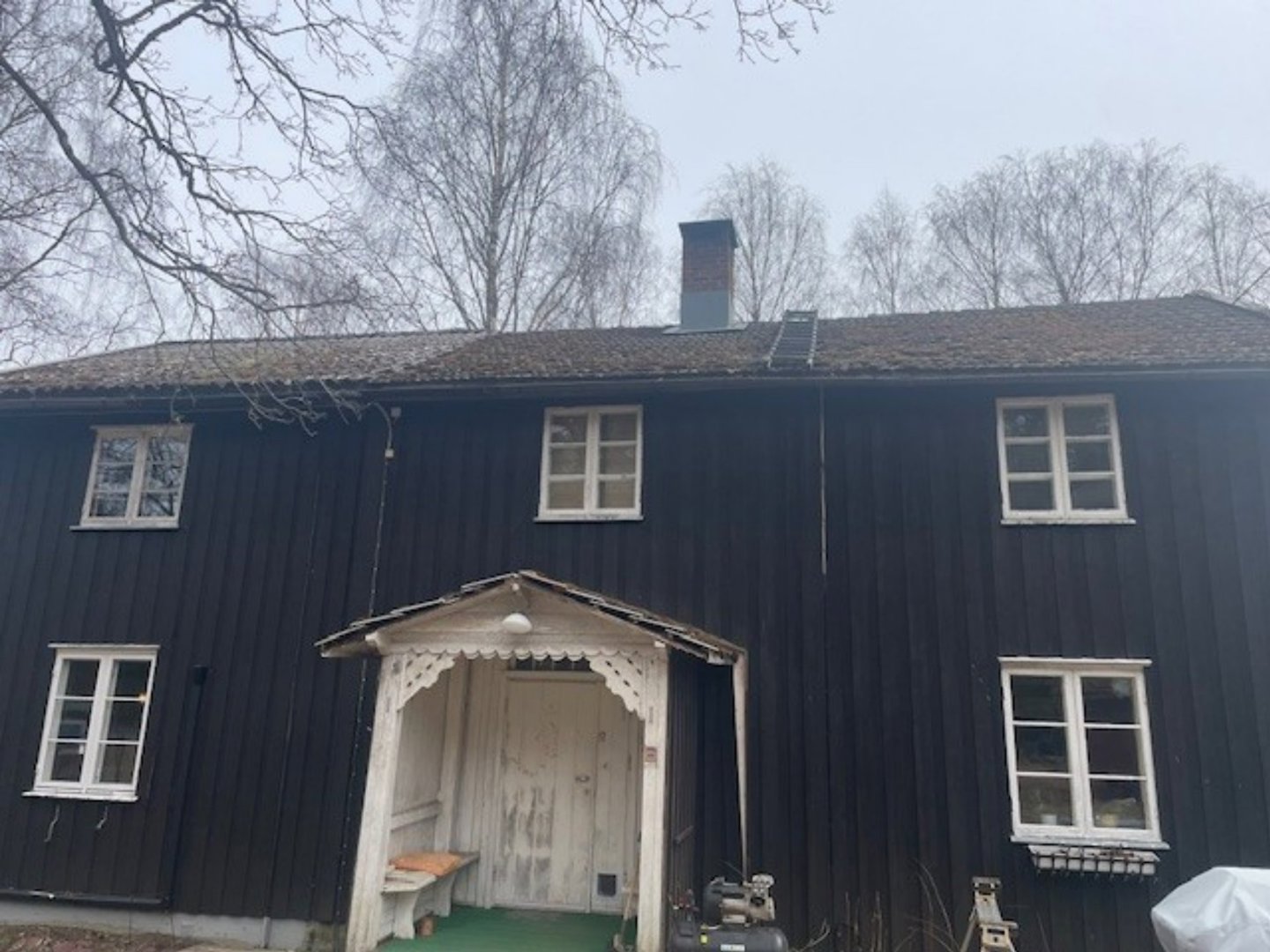 Fotografi av en mørk, trolig svart, trebygning sett fra fronten. Bygningen har hvite vinduer, en hvit inngangsparti med dekorativt tak, og en røykstak. Bakgrunnen består av bar trær og en grå himmel.