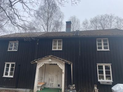 Fotografi av en mørk, trolig svart, trebygning sett fra fronten. Bygningen har hvite vinduer, en hvit inngangsparti med dekorativt tak, og en røykstak. Bakgrunnen består av bar trær og en grå himmel.