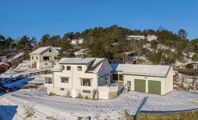 Fotografi av en hvit villa med grønn garasjeport, sett fra lufta. Huset er omgitt av snø og ligger i et boligområde med skogbakke.