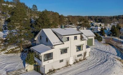 Fotografi av en hvit villa med grønn garasjeport, sett fra lufta. Huset er belagt med snø og omgitt av skog og nabolag.