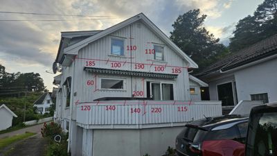 Fotografi av en hvit, trepanelert hytte med overlagte røde målelinjer og dimensjoner (f.eks. 185, 100, 190) som indikerer en måling av fasadens bredde og vindusposisjon.