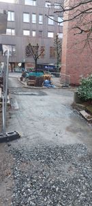 Fotografi av et byggeplassområde med en nybetont vei eller gangvei i forgrunnen, omgitt av byggematerialer og en eksisterende bygning i bakgrunnen.