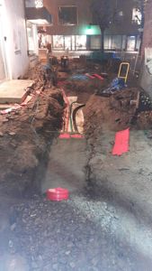 Fotografi av en utgravet grøft med rør og materialer under et bygg eller i en gate.