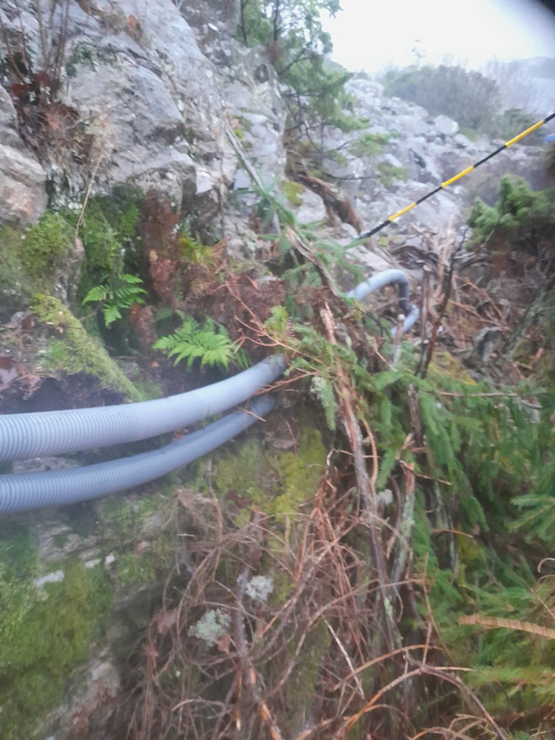 Fotografi av en grå, ribbet rørledning (sannsynligvis for vann eller avløp) som er lagt langs en steinvegg i et naturlig landskap med mose og busker.