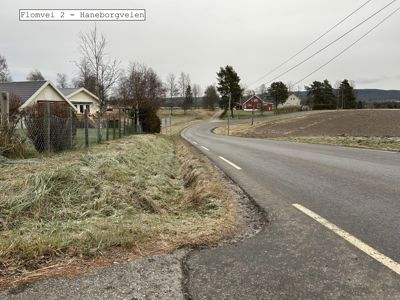 Fotografi av en vei (Flomvei 2 - Haneborgveien) med hus og landbruksarealer i bakgrunnen.