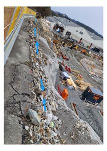 Fotografi av et byggeplassområde med en steinete skrent, en gul sikkerhetshekk og en hvit industribygning i bakgrunnen.