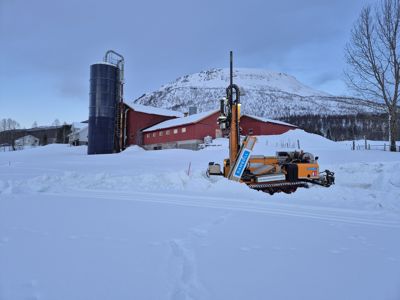 Fotografi av en byggeplass i snø med boremaskin, silo og rød bygning i bakgrunnen.