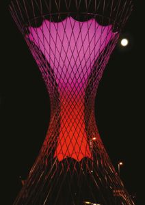 Fotografi av en lysende, nettkonstruksjon (trolltårn) mot nattlig himmel med måne.