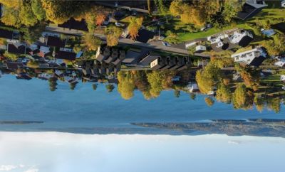 Luftfoto av et boligområde med hus, veier og trær, sett mot en stor vannflate og fjell i bakgrunnen.