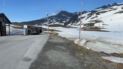 Fotografi av en vei i fjellområde med snø, en bil og en liten bygning.