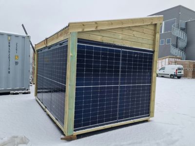 Fotografi av en ferdigstilt eller under bygging solcelleanlegg (solar shelter) med solpaneler, sett fra siden. Bakgrunnen viser en container og en annen bygning i snø.