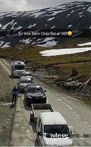 Fotografi av en kø av biler på en vei i fjellområde med snø. Bildet inneholder også en tekstoverlegning og et vannmerke.