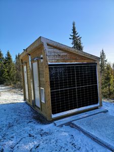 Fotografi av en liten, trekonstruert bygning (kanskje en utedo eller teknisk rom) med solpaneler på siden, sett i et snødekt landskap.