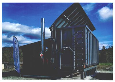 Fotografi av en moderne, mørk bygning med et markant tak og en høy røykledning, sett fra utsiden.