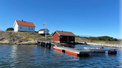 Fotografi av en bygning (hvit med rødt tak) og en mindre rød uthus ved vannkanten, sett fra et vassdriftspunkt.