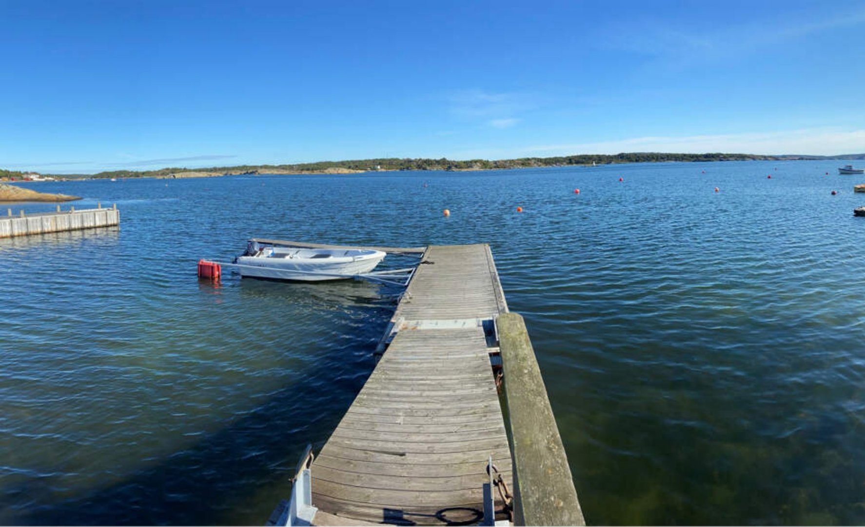 Fotografi av en kai eller brygge som strekker seg ut i vannet, med en båt ved siden av. I bakgrunnen er det land og en klar himmel.