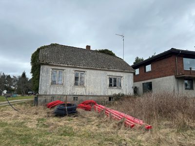 Fotografi av en gammel, hvitmalt trehytte med etasjehus i bakgrunnen. Foran ligger røde og svarte rør på grunnen.