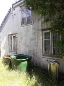 Fotografi av en eksisterende, hvitmalt trebygning sett fra siden. Bildet viser fasaden med vinduer, takkant, og grønt i forgrunnen med søppelcontainere.