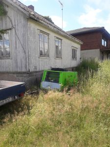 Fotografi av en gammel, hvitfarget trehytte med etasjeforlengelse, sett fra siden. I bakgrunnen er det en annen bygning (bricks), og i forgrunnen står det en grønn maskin og en trailer.