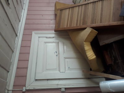 Fotografi av en hvit dør med paneler, innrammet i et hvitt karmverk på en rosa fasade. Bildet viser også deler av en trapp og byggematerialer.